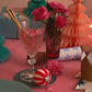 A decorated table with Peppermint Candy Surprise Balls by Meri Meri, a vase of pink roses, wrapped gifts, party crackers, a crown, and festive decor on a pink cloth—perfect for holiday surprises or stocking fillers.