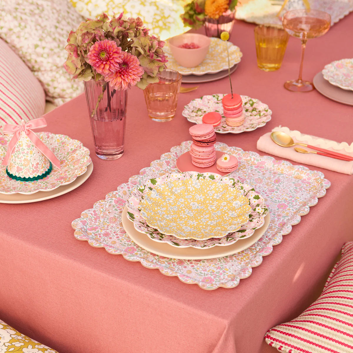 A pink tablecloth with floral plates, glasses, napkins, a vase of flowers, macarons, and decorative pillows arranged on the charming scalloped-edge Wharfedale Tray by Meri Meri.