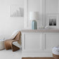 Minimalist room with a wavy rattan chair, white cabinet, Visual Comfort Melanie Large Table Lamp Collection in reactive ceramic glazes with a silk shade, framed black-and-white photos, woven basket, and beige and white neutral decor.