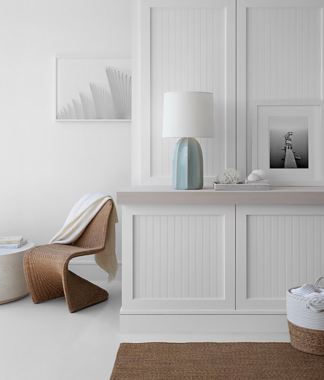 Minimalist room with a wavy rattan chair, white cabinet, Visual Comfort Melanie Large Table Lamp Collection in reactive ceramic glazes with a silk shade, framed black-and-white photos, woven basket, and beige and white neutral decor.