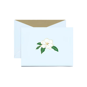 The Magnolia Blossom Classic Card Set by Crane features a light blue card with a white magnolia and green leaves, plus a matching envelope with gold interior—perfect for sending heartfelt notes symbolizing strength.