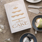 A white PrintWorks "The Essentials Cake Tools" boxed set sits on a table with cake servers, plates of cake, forks, and champagne glasses—both stylish kitchen decor and the perfect tools for cake serving.