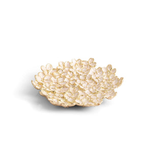 The Michael Aram Cherry Blossom Collection features a decorative bowl with a textured surface that evokes soft pink Japanese flower petals on a white background.