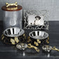 Two metal dog bowls with gold butterfly accents from the Michael Aram Flights of Fancy Collection, a coordinating treat jar with a bone-shaped handle, two small hand-sculpted clay bowls, and a framed photo of a dog—along with the Dog Scoop—create whimsical decor on a dark surface.
