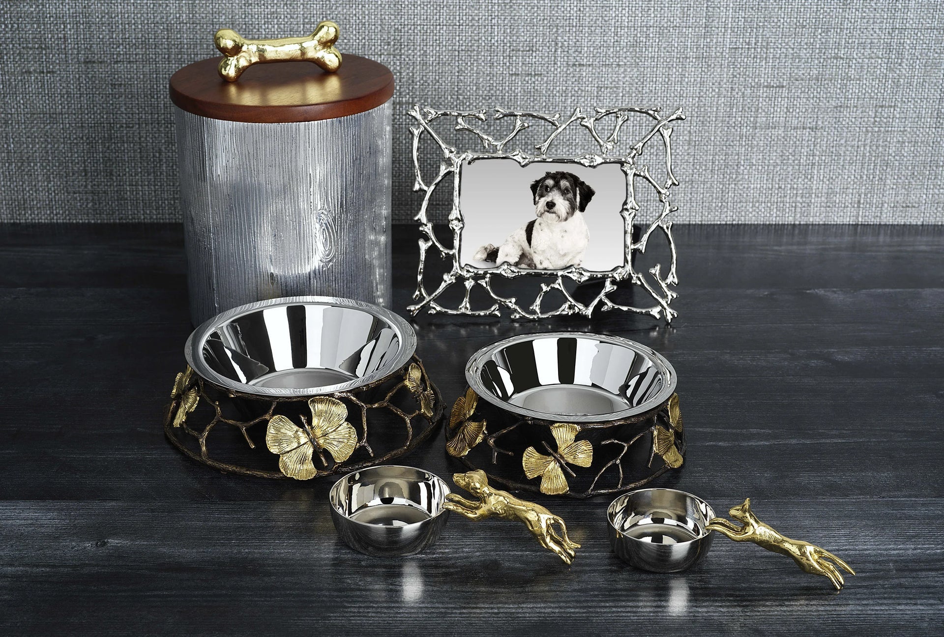 Two metal dog bowls with gold butterfly accents from the Michael Aram Flights of Fancy Collection, a coordinating treat jar with a bone-shaped handle, two small hand-sculpted clay bowls, and a framed photo of a dog—along with the Dog Scoop—create whimsical decor on a dark surface.