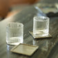 Two textured glass tumblers from Michael Aram’s Ivy & Oak Glass and Coaster Gift Set, one on a square coaster and the other beside an empty one, display rustic elegance; a tray from the set appears in the blurred background.
