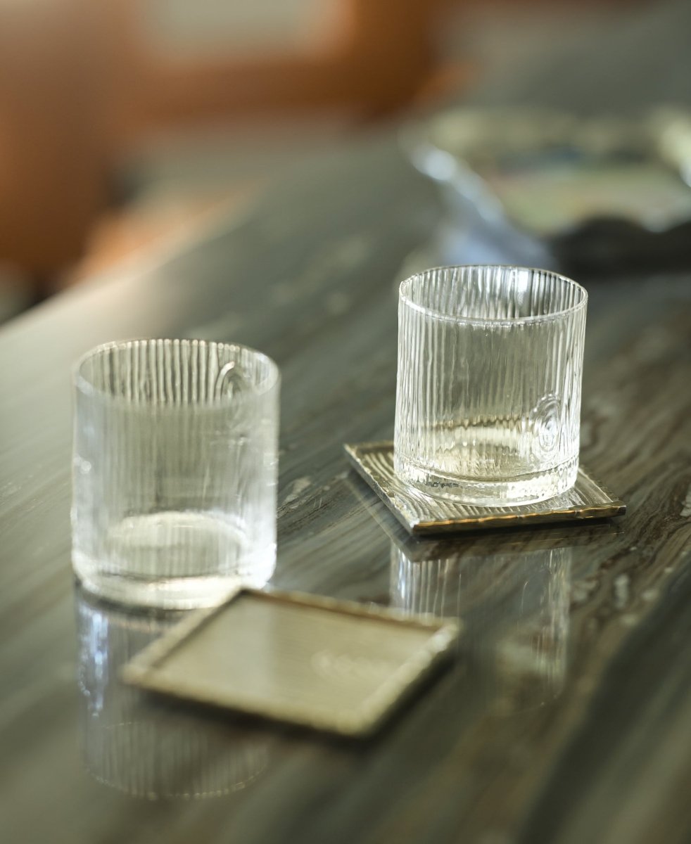 Two textured glass tumblers from Michael Aram’s Ivy & Oak Glass and Coaster Gift Set, one on a square coaster and the other beside an empty one, display rustic elegance; a tray from the set appears in the blurred background.