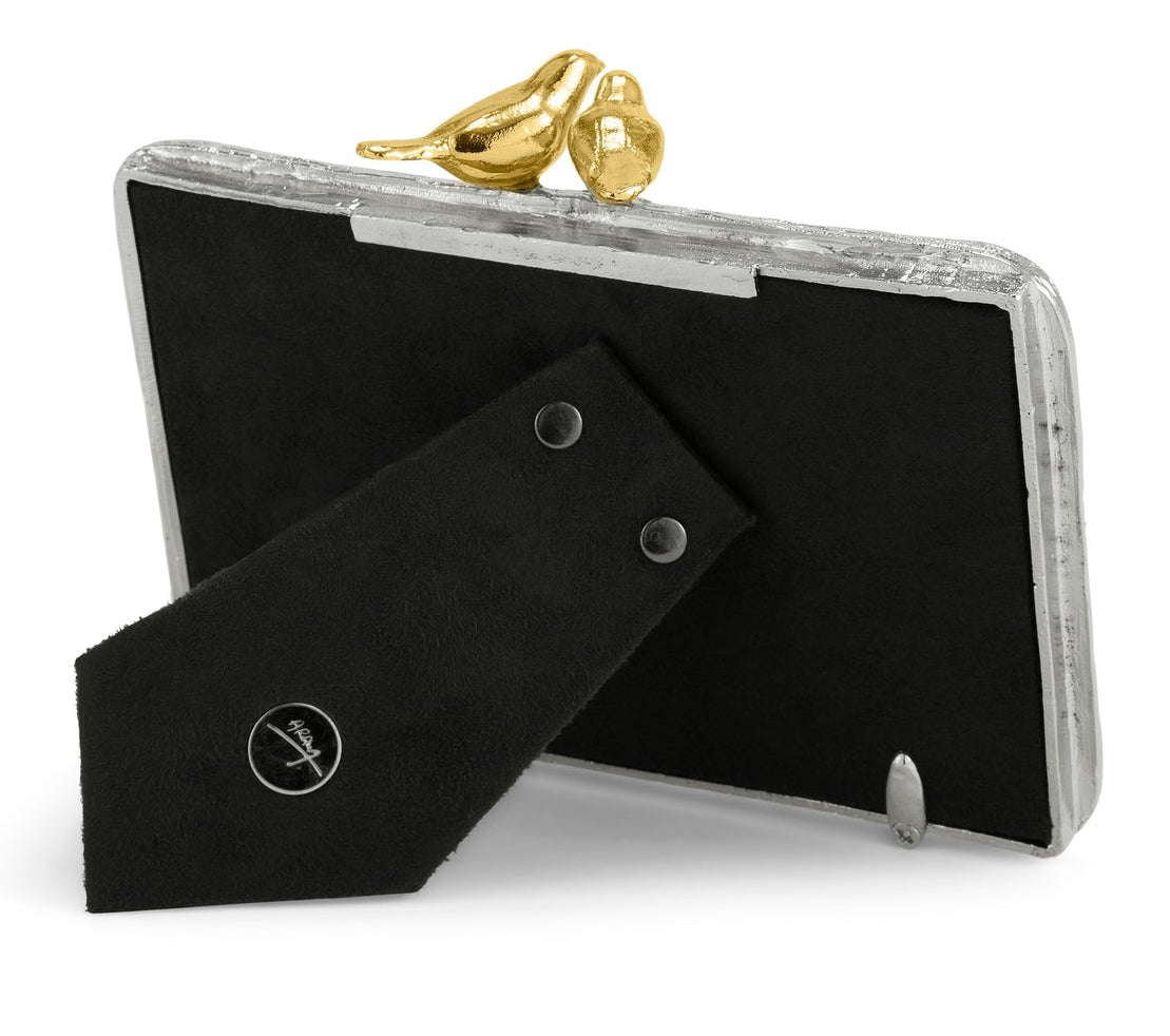 The Michael Aram Lovebirds Frame is a handcrafted picture frame featuring silver edges, a black back, a stand, and two gold bird accents on top.