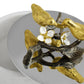 A close-up of the Michael Aram Lovebirds Trinket Box, featuring two gold-tone birds on a branch with white flowers, displayed on a reflective surface.