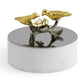 The Michael Aram Lovebirds Trinket Box is a round silver box topped with a sculpted Tree of Life branch, gold-toned birds, and glass flowers—an elegant way to store jewelry.