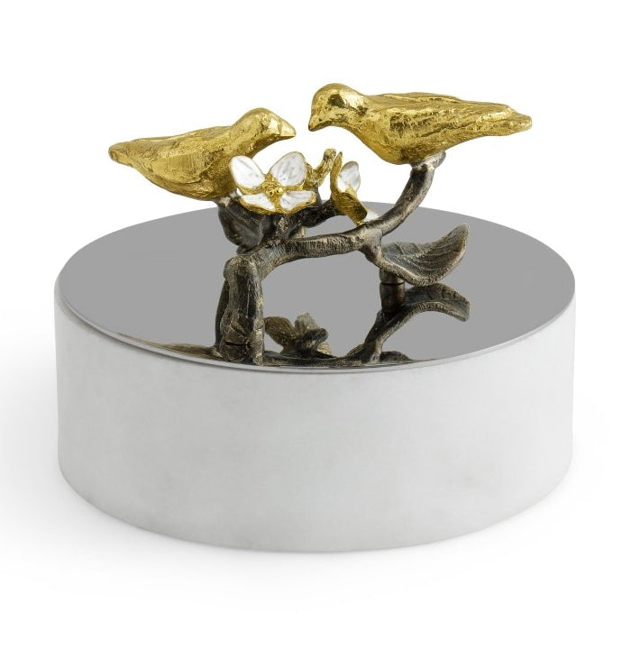 The Michael Aram Lovebirds Trinket Box is a round silver box topped with a sculpted Tree of Life branch, gold-toned birds, and glass flowers—an elegant way to store jewelry.