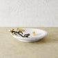 The Michael Aram Lovebirds Trinket Dish features a white marble base with a gold and bronze bird and branch accent on the rim, holding a single gold object. It sits on a beige surface against a textured wall.