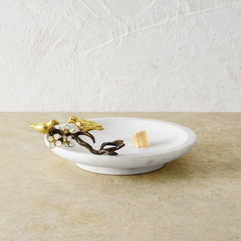The Michael Aram Lovebirds Trinket Dish features a white marble base with a gold and bronze bird and branch accent on the rim, holding a single gold object. It sits on a beige surface against a textured wall.