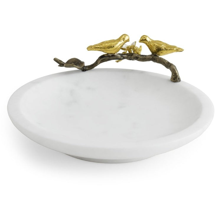 The Lovebirds Trinket Dish by Michael Aram is a white, round jewelry holder inspired by the Tree of Life, featuring a decorative gold branch on the rim with two gold bird figures facing each other.