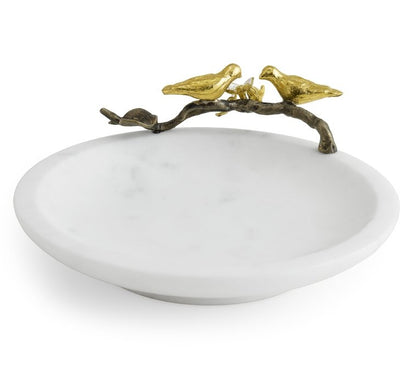 The Lovebirds Trinket Dish by Michael Aram is a white, round jewelry holder inspired by the Tree of Life, featuring a decorative gold branch on the rim with two gold bird figures facing each other.