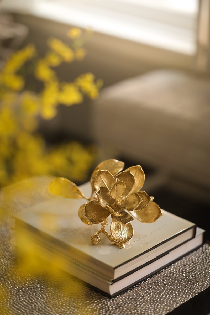 A handcrafted brass Magnolia Decorative Object by Michael Aram sits atop two stacked hardcover books, softly lit with blurred yellow flowers in the background.