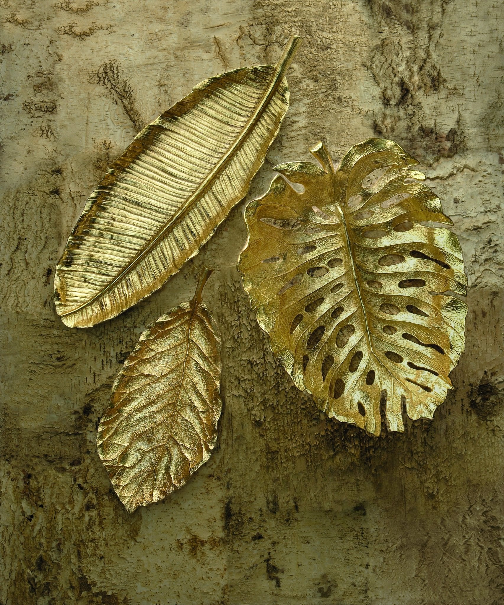 Three metallic gold leaves in varying shapes from the Michael Aram New Leaves Serveware Collection rest on a textured brown surface—handcrafted, nature-inspired decor blending artistry and function.