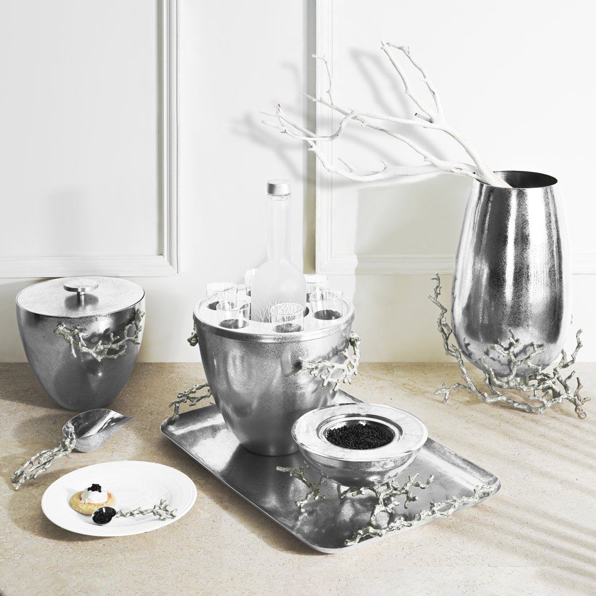 A metallic tabletop display showcases Michael Aram’s Ocean Reef Serveware and Barware Collection with a silver ice bucket, matching bowls, a vase with white branches, a tray, and hors d’oeuvres—all set against a white wall.