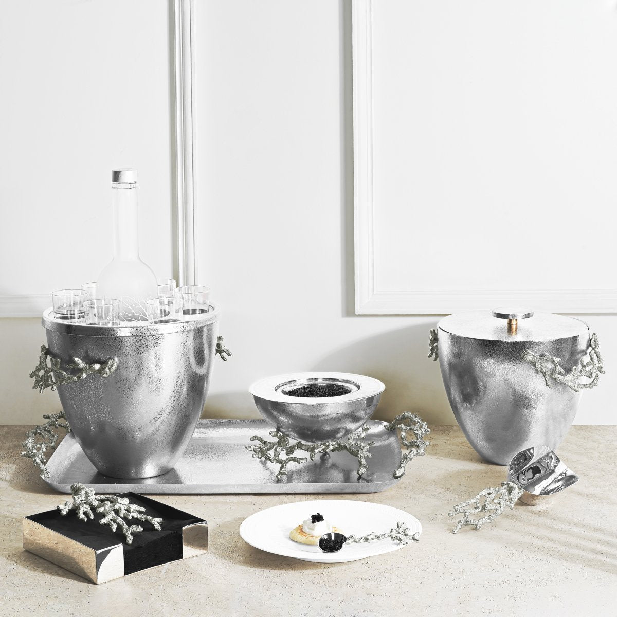 A countertop displays Michael Aram’s Ocean Reef Serveware and Barware Collection, featuring a silver set with branch-like handles, serving bowls, glasses, trays, containers, and a dessert plate.
