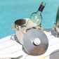 A wine bottle rests in the Michael Aram Ocean Reef Ice Bucket, its stainless steel design and nearby lid adding elegance to a poolside table.