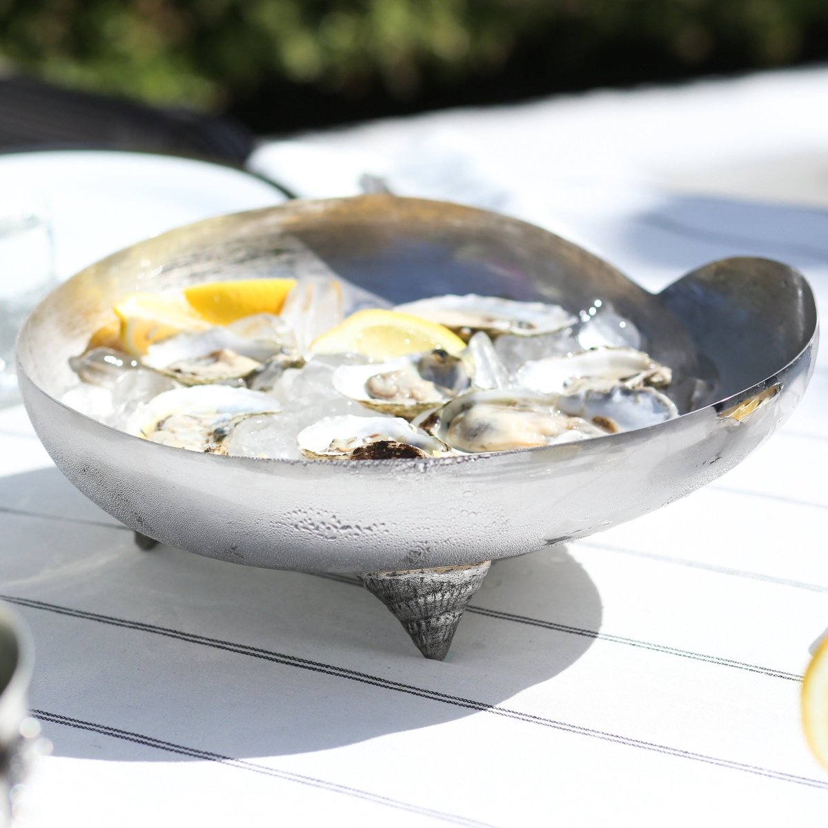 A metal serving bowl from the Michael Aram Ocean Reef Serveware and Barware Collection holds ice, opened oysters, and lemon wedges atop a striped white tablecloth outdoors, capturing the vibrant freshness of coral reefs.