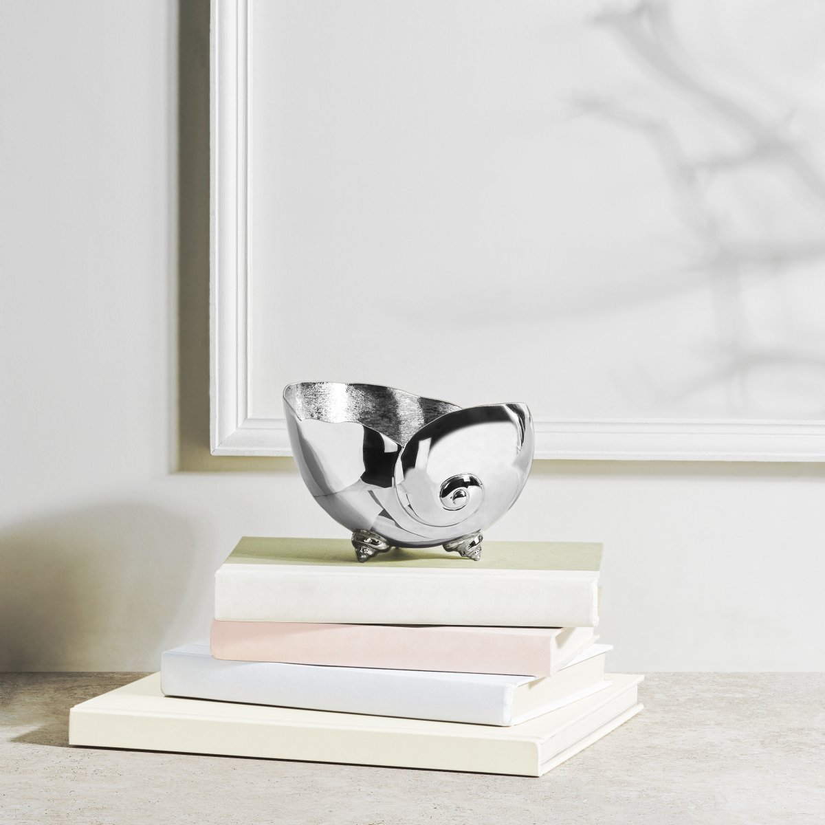 A shiny silver nautilus shell bowl from the Michael Aram Ocean Reef Serveware and Barware Collection sits atop four light-covered books against a white wall—an ideal accent for coral reef decor lovers.
