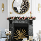 The Palm Decorative Fireplace Screen by Michael Aram sits before a black marble fireplace with antique gold accents, adorned for the holidays with garland and red ribbon. A mirror above reflects a decorated tree, surrounded by luxury furnishings and gifts.