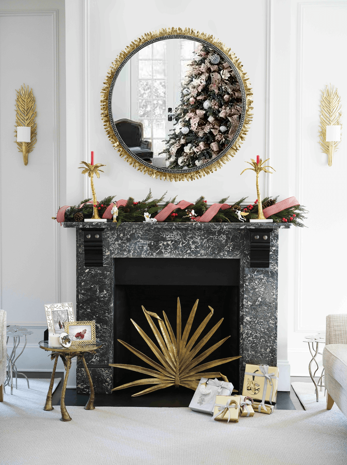 The Palm Decorative Fireplace Screen by Michael Aram sits before a black marble fireplace with antique gold accents, adorned for the holidays with garland and red ribbon. A mirror above reflects a decorated tree, surrounded by luxury furnishings and gifts.