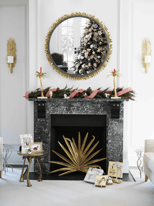 The Palm Decorative Fireplace Screen by Michael Aram sits before a black marble fireplace with antique gold accents, adorned for the holidays with garland and red ribbon. A mirror above reflects a decorated tree, surrounded by luxury furnishings and gifts.