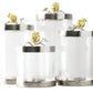 Four clear textured glass Pomegranate Silver & Gold Canisters by Michael Aram, each with a metallic lid topped by a gold pomegranate and leaf accent—symbols of abundance—displayed against a white background.