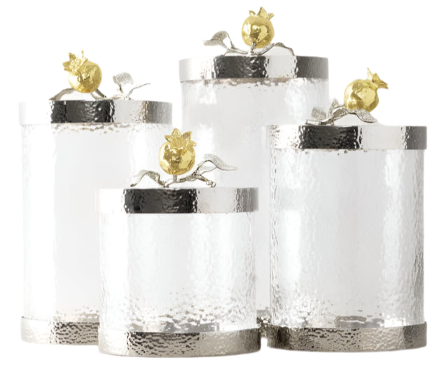 Four clear textured glass Pomegranate Silver & Gold Canisters by Michael Aram, each with a metallic lid topped by a gold pomegranate and leaf accent—symbols of abundance—displayed against a white background.