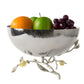A bowl from the Michael Aram Pomegranate Silver & Gold Serveware Collection holds an orange, a green apple, and red grapes. Its base has branch-like details with elegant leaves. Crafted in stainless steel and nickelplate.