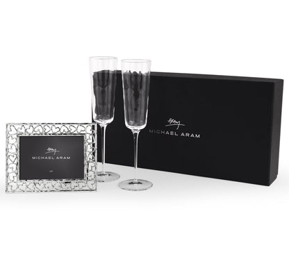 The Michael Aram Reflective Champagne Flute & Heart Frame Gift Set features two flutes and a polished photo frame, elegantly displayed in front of a black Michael Aram box.