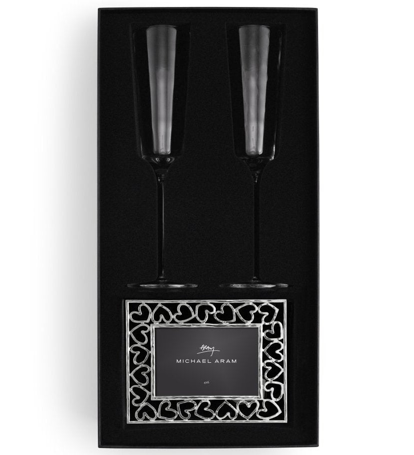 The Michael Aram Reflective Champagne Flute & Heart Frame Gift Set includes two reflective champagne flutes and a decorative stainless steel photo frame with heart-shaped accents.