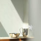 A polished stainless steel bowl and pitcher from Michael Aram’s Reflective Serveware Collection sit on a wooden table in a sunlit room, with soft light and shadows enhancing the calm, modern vibe.