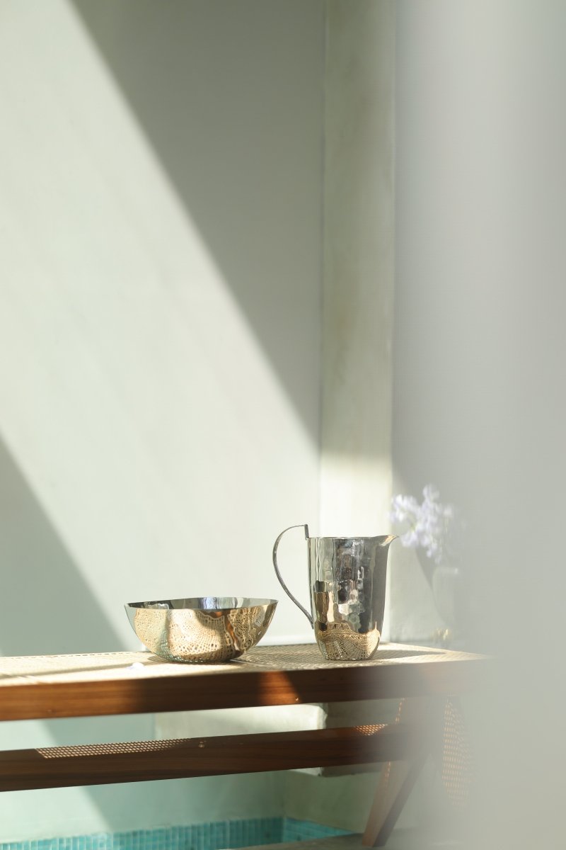 A polished stainless steel bowl and pitcher from Michael Aram’s Reflective Serveware Collection sit on a wooden table in a sunlit room, with soft light and shadows enhancing the calm, modern vibe.