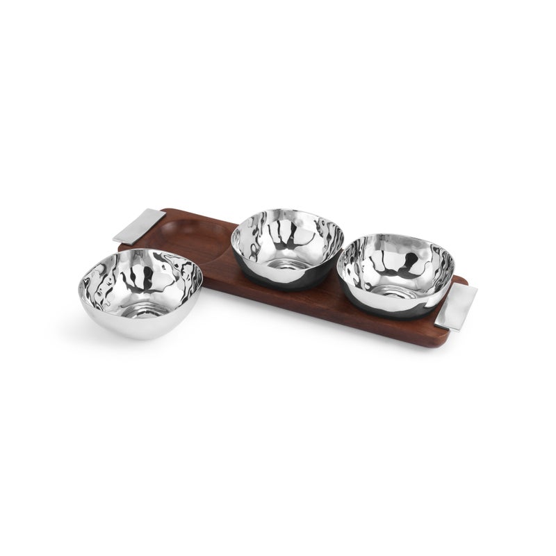 Four hammered metal bowls from the Michael Aram Reflective Serveware Collection rest on a rectangular wooden tray with metal handles; one highly polished stainless steel bowl is placed beside the tray for a modern touch.