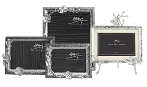 The White Orchid Frame Collection by Michael Aram features four silver photo frames with graceful orchid motifs; three are upright, and one is displayed on a decorative stand.