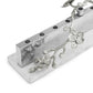 The Michael Aram White Orchid Judaica Collection Menorah features a silver, hammered metal design with floral branch and blossom accents on a white rectangular base.