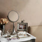A vanity with a round mirror, silver decorative items, a vase of roses, a framed photo, jewelry displayed on the Michael Aram White Orchid Ring Catchall, and a beige sofa in the background.