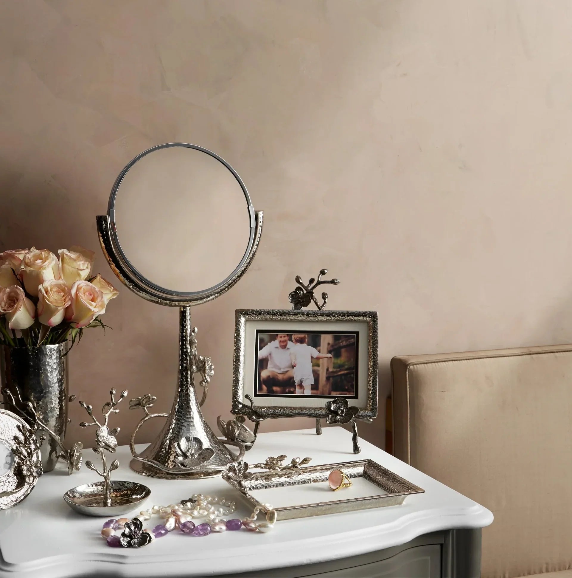 A vanity with a round mirror, silver decorative items, a vase of roses, a framed photo, jewelry displayed on the Michael Aram White Orchid Ring Catchall, and a beige sofa in the background.