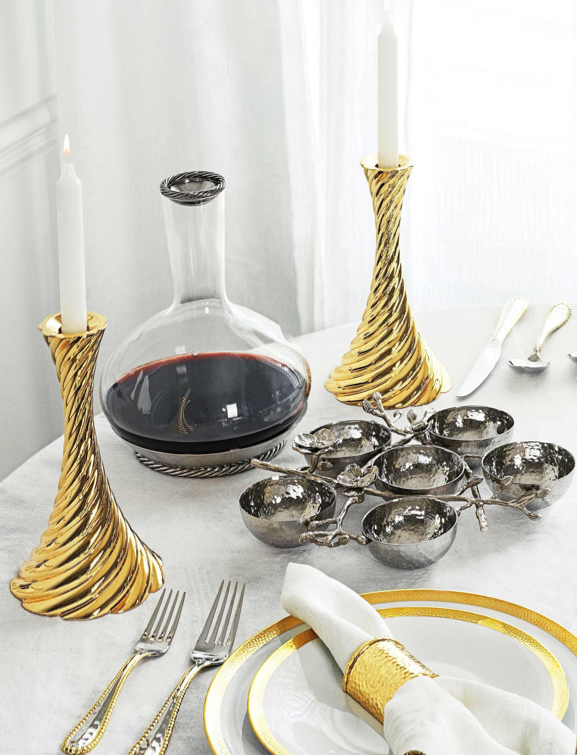 A formal table set with Michael Aram’s White Orchid Judaica Collection, featuring gold-rimmed plates, a Seder Plate, glass decanter, metal serving bowls, gold and silver candlesticks, and elegant cutlery on a white cloth.