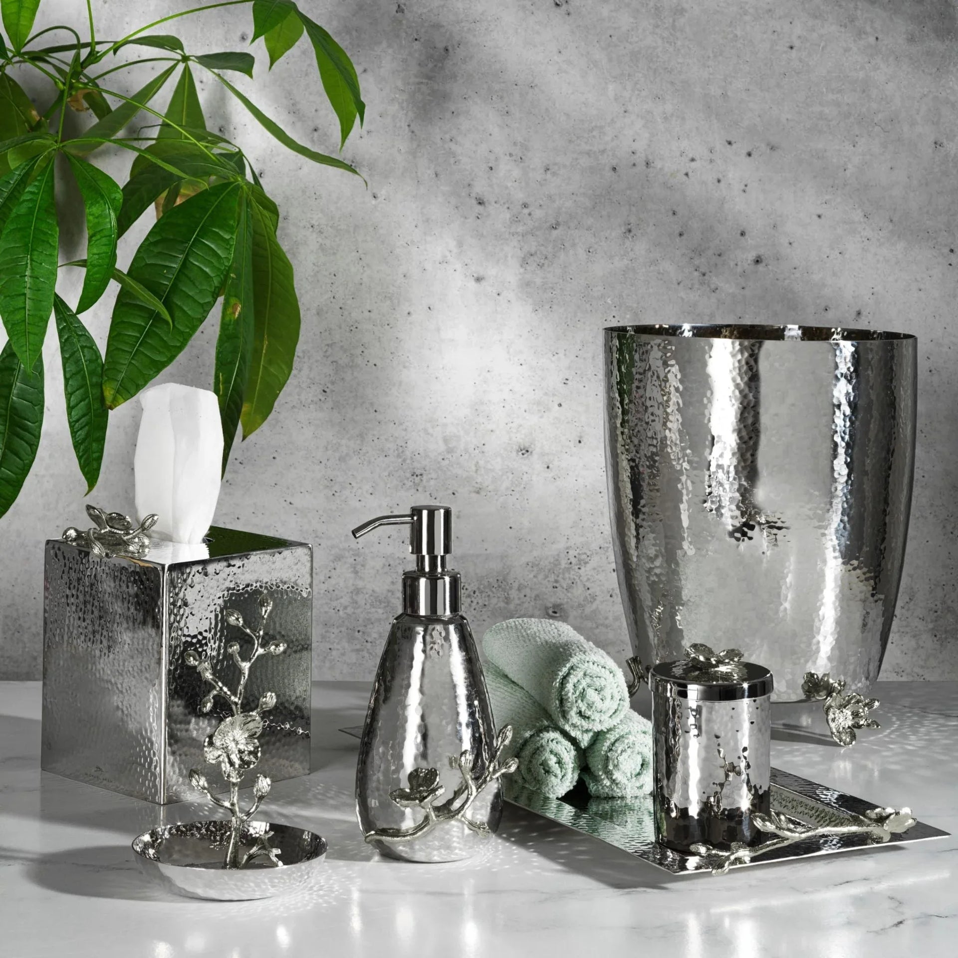The Michael Aram White Orchid Bath Collection, featuring luxury bathroom accessories such as a soap dispenser, tissue box, waste bin, tumbler, tray, and dish in a silver hammered metal design, is elegantly displayed on a marble surface with a green plant nearby.