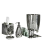 The Michael Aram White Orchid Bath Collection is a six-piece bathroom accessory set featuring a luxurious hammered texture in silver, with elegant pieces including a soap dispenser, tissue box, tumbler, soap dish, waste bin, and rolled green towels.