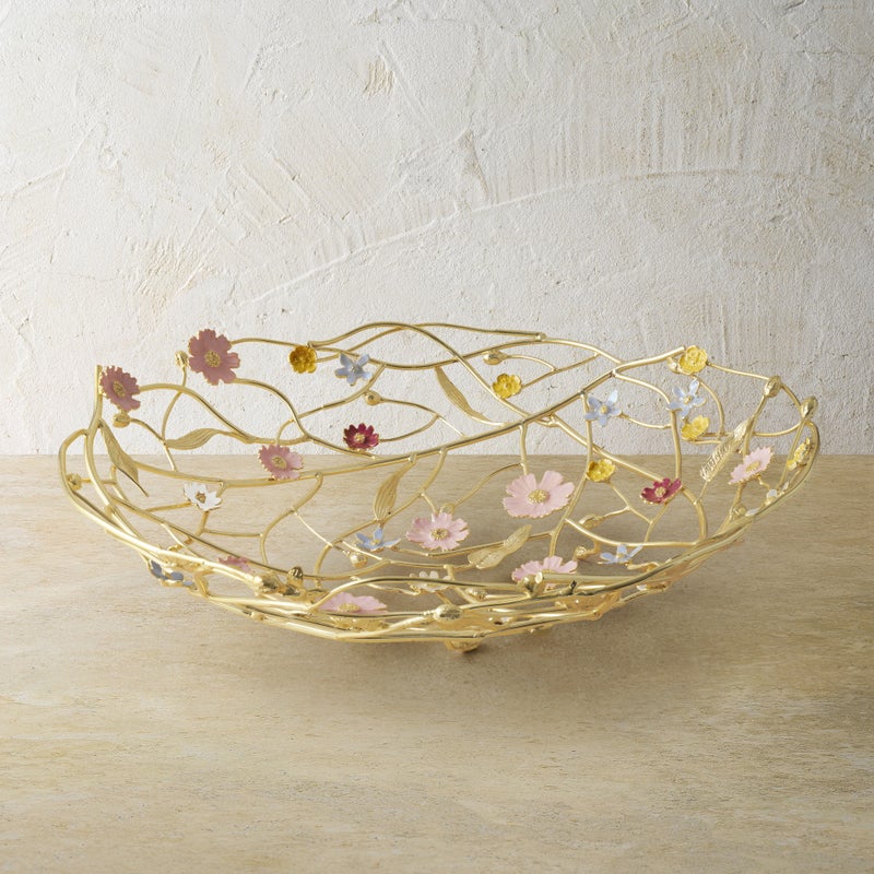 The Michael Aram Wildflower Serveware Collection features a gold wire centerpiece basket with vibrant, handcrafted wildflowers, displayed on a light beige surface against a textured wall.