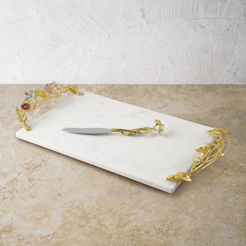 From the Wildflower Serveware Collection by Michael Aram, a rectangular white marble cheeseboard with ornate gold handles and handcrafted wildflowers comes with a matching gold and silver knife, shown on a beige stone surface.