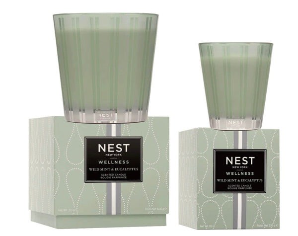 Two green glass candles labeled "NEST Wild Mint & Eucalyptus Candle Collection" are placed on matching printed boxes, one larger than the other. The invigorating blend of wild mint and eucalyptus promises a refreshing escape with every burn.