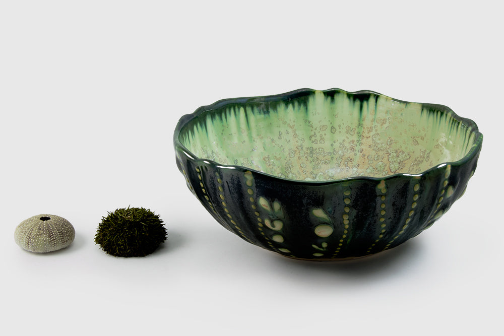 From AE Ceramics’ Sea Urchin Bowl Collection, a handmade green ceramic bowl with a textured, scalloped rim sits beside a sea urchin shell and mossy sphere on a white background.