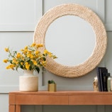 The Amani Round Abaca Rope Mirror by Made Goods hangs on a light wall above a wooden table adorned with a vase of yellow flowers, books, and a small decorative item.