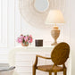 A white desk with a vase of pink flowers, a black box, and a woven lamp sits beneath the Made Goods Saffron Mirror. A gold chair in front enhances the retro vibe.